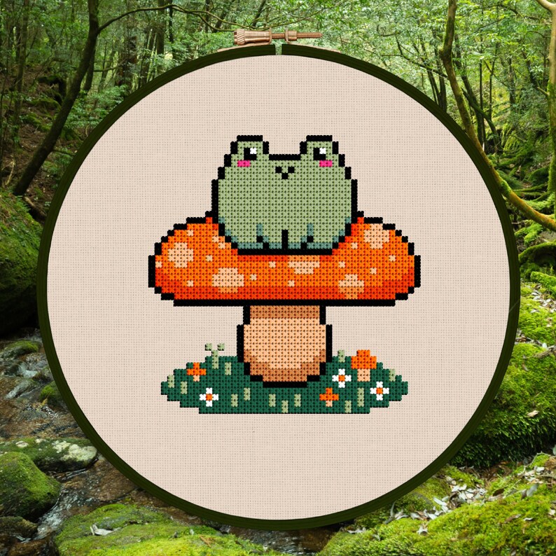 May include: Cross-stitch embroidery of a green frog sitting on a red and orange mushroom. The mushroom is surrounded by green grass and small white flowers. The embroidery is framed in a wooden hoop.