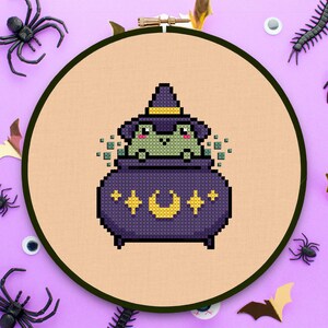 May include: A green frog wearing a witch's hat sits inside a purple cauldron with a yellow crescent moon and stars. The cauldron is surrounded by a light orange background.