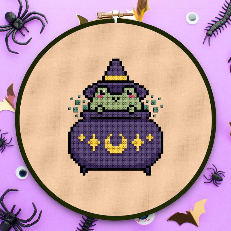 May include: A green frog wearing a witch's hat sits inside a purple cauldron with a yellow crescent moon and stars. The cauldron is surrounded by a light orange background.