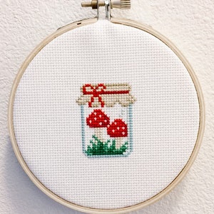 May include: A white embroidery hoop with a cross-stitch design of a glass jar filled with red mushrooms and green grass. The jar has a brown lid with a red bow.