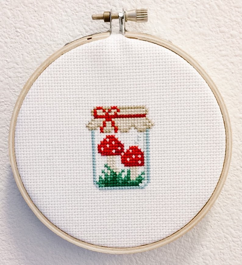 May include: A white embroidery hoop with a cross-stitch design of a glass jar filled with red mushrooms and green grass. The jar has a brown lid with a red bow.