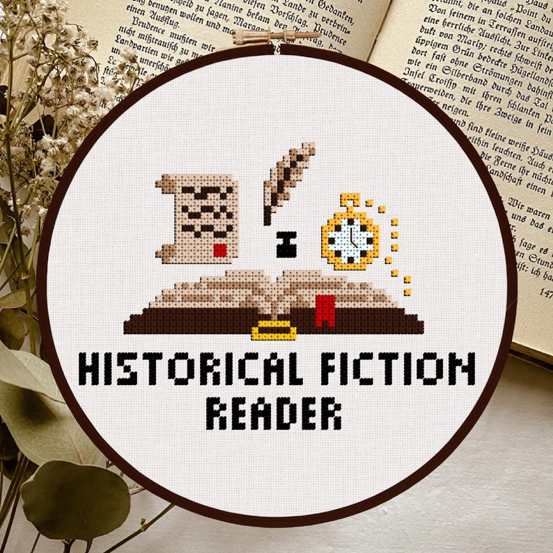 May include: A cross-stitch embroidery hoop featuring a design for a historical fiction reader. The design includes an open book, a scroll, a quill, and a pocket watch. The text "HISTORICAL FICTION READER" is stitched below the design.