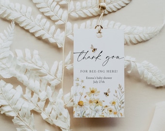 May include: A white card with a gold tassel attached to the top. The card has a floral design with bees and the text "Thank you for bee-ing here! Emma's baby shower July 27th".