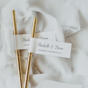 Puede incluir: Pajitas de papel doradas con etiquetas blancas con nombres. Las etiquetas dicen "Michelle & Brian" y "September 19 • San Francisco, CA". Las pajitas están sobre un fondo de tela blanca suave.