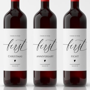 Puede incluir: Tres botellas de vino con etiquetas blancas que dicen "Cheers to your first" seguido de "Christmas", "Anniversary" y "Fight". Cada etiqueta tiene un pequeño corazón y los nombres Celeste & Robert y la fecha 21, 23 o 25 de agosto.