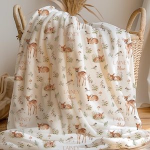 May include: A cream-coloured baby blanket with a woodland theme, featuring deer, mushrooms, and floral elements. The blanket is personalised with the name "Lainey Mae" in a soft brown font. The blanket is folded in a wicker basket.
