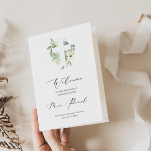 May include: A white card with a watercolour illustration of a cross surrounded by green leaves and white flowers. The text on the card reads "Welcome to the baptism of our daughter Mia Pearl St. Cecilia Catholic Church August 27, 20XX".