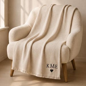 May include: A cream-colored waffle-knit throw blanket with fringe edges, featuring the black embroidered letters KME and a small black heart symbol positioned near the bottom corner of the fabric, draped over a matching cream-colored armchair.