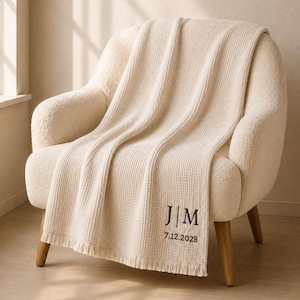 May include: A cream-colored waffle-knit throw blanket with fringe edges, featuring the embroidered black text "J|M" and the date "7.12.2028" in the bottom corner. The blanket is draped over a textured, off-white upholstered armchair with wooden legs.