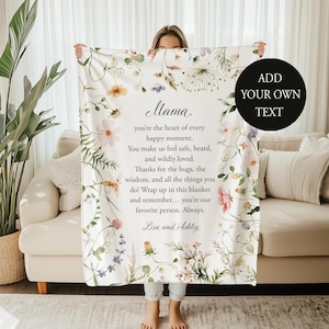 May include: A white blanket with a floral design and the text "Mama" in elegant script. The blanket is adorned with colorful flowers and greenery. The text reads, "you're the heart of every happy moment...you're our favorite person. Always." A black circle with the text "ADD YOUR OWN TEXT" is on the right.