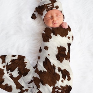 May include: A newborn baby wrapped in a cowhide print swaddle blanket and matching hat. The hat and blanket feature brown and white spots. The name "Wyatt Austin" is printed on a brown patch on the hat and blanket.