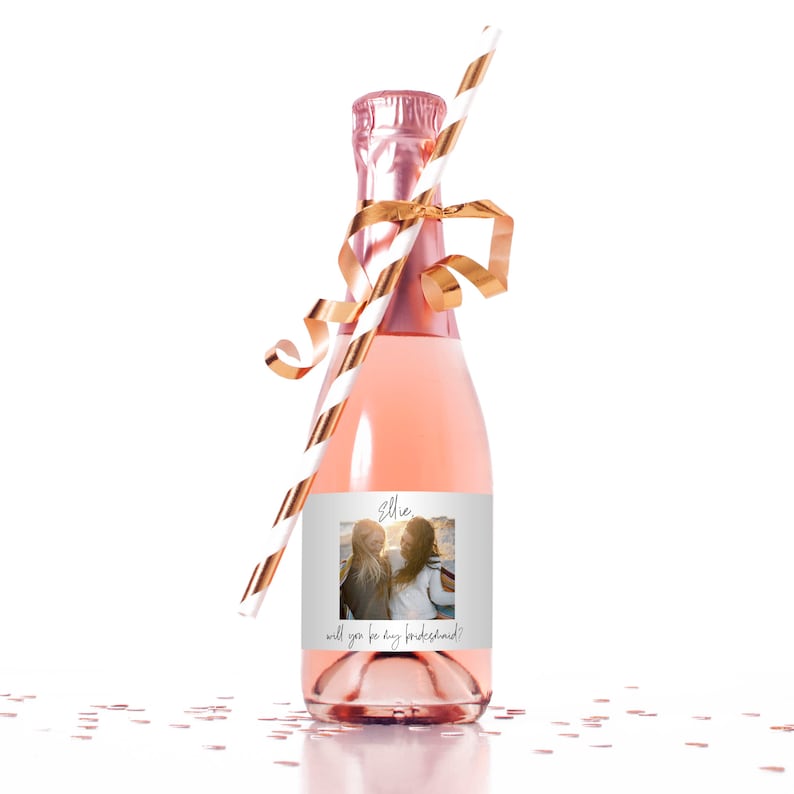 Peut inclure: Une petite bouteille de champagne ros&eacute; avec un ruban dor&eacute; et une paille ray&eacute;e. La bouteille porte une &eacute;tiquette avec une photo de deux femmes et le texte "Ellie, veux-tu &ecirc;tre ma demoiselle d'honneur ?"