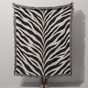May include: A rectangular woven throw blanket featuring a high-contrast black and off-white zebra stripe pattern. The blanket is finished with decorative fringe along all four edges, creating a textured border around the animal print design.