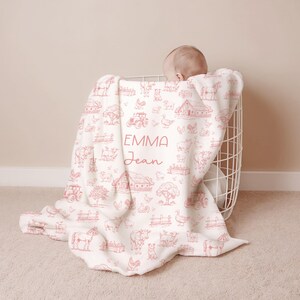 May include: A white baby blanket featuring a repeating red line-art pattern of farm animals, tractors, and barns. The center of the blanket displays the name EMMA Jean in a combination of block and script lettering in red ink.