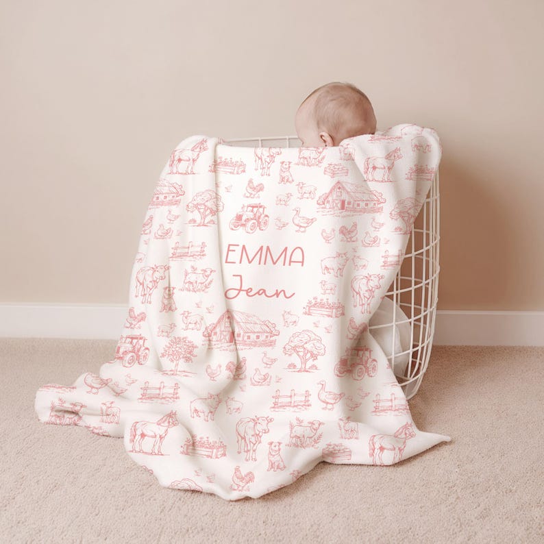 May include: A white baby blanket featuring a repeating red line-art pattern of farm animals, tractors, and barns. The center of the blanket displays the name EMMA Jean in a combination of block and script lettering in red ink.