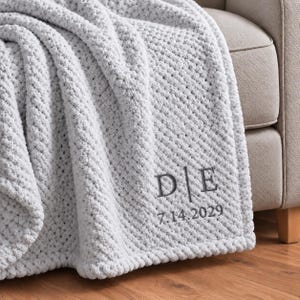 以下が含まれることがあります： ライトグレーのテクスチャードニットスローブランケット。パーソナライズされた刺繍デザインが施されています。刺繍には縦線で区切られたDとEの文字があり、その下にダークグレーの糸で7.14.2029という日付が縫い付けられています。