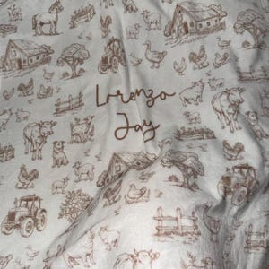 May include: A white fabric blanket featuring a repeating brown line-art pattern of farm animals, barns, tractors, and trees. The center of the blanket displays the name Lorenzo Jay printed in a brown cursive script font.