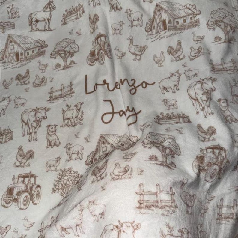 May include: A white fabric blanket featuring a repeating brown line-art pattern of farm animals, barns, tractors, and trees. The center of the blanket displays the name Lorenzo Jay printed in a brown cursive script font.