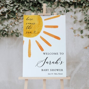 May include: A white welcome sign with a yellow and orange watercolor sun design. The text reads "Here comes the sun" and "Welcome to Sarah's Baby Shower June 18 - Oahu, Hawaii".