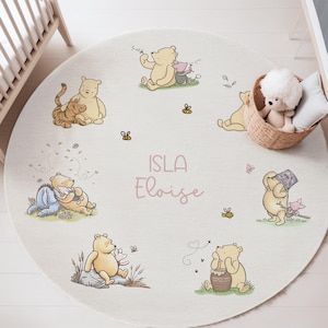 May include: A circular rug with a Winnie the Pooh theme, featuring Pooh, Piglet, Tigger, and Eeyore illustrations. The rug is off-white with the name "Isla Eloise" in pink. Bees and butterflies are scattered around the characters.