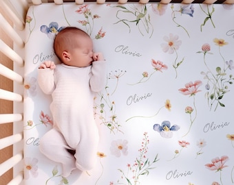 Puede incluir: Un bebé con un body blanco está acostado sobre una sábana. La sábana es blanca con un estampado floral de flores rosas, azules y amarillas. El nombre "Olivia" está impreso en la sábana en letra cursiva.