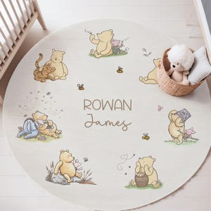 May include: A circular, cream-coloured nursery rug with Winnie the Pooh characters. The rug features illustrations of Pooh, Piglet, Eeyore, and Tigger, along with bees and the names "Rowan James". A wicker basket with a stuffed toy and pillow is in the background.