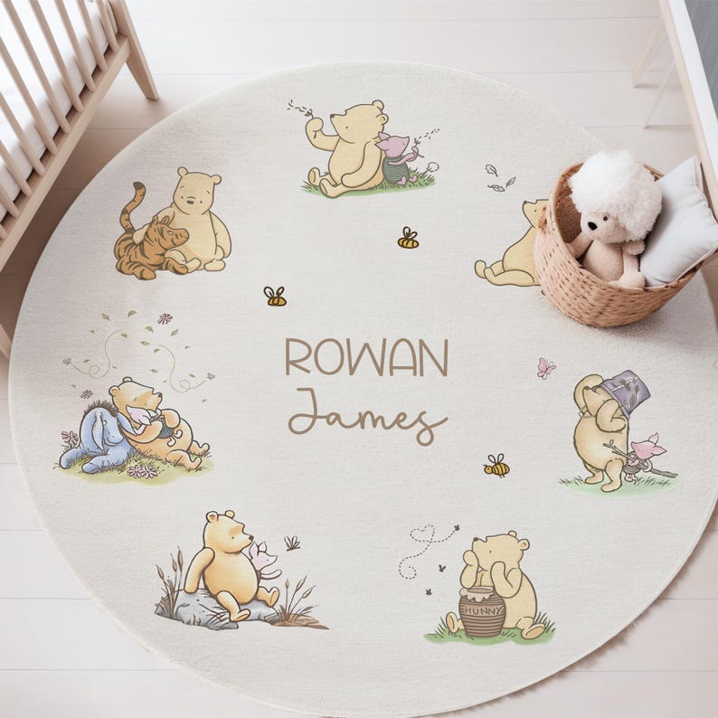 May include: A circular, cream-coloured nursery rug with Winnie the Pooh characters. The rug features illustrations of Pooh, Piglet, Eeyore, and Tigger, along with bees and the names "Rowan James". A wicker basket with a stuffed toy and pillow is in the background.