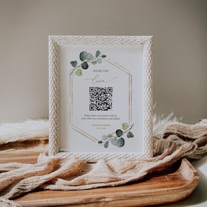 May include: White frame with a gold hexagon and green leaves. The text "SHARE THE love" is written in cursive. A QR code is in the center of the hexagon. The text below the QR code reads "Please share your photos with us. Scan with your smartphone and upload. Heather & Michael 10.07.202X"