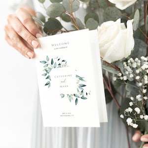 May include: A white wedding program with a green leafy wreath design. The text reads "Welcome to the wedding of Catherine and Mark, July 12, 2022, San Francisco, CA."