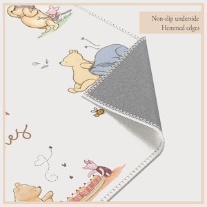 May include: A rectangular rug with illustrations of Winnie the Pooh and friends. The rug has a white background, stitched border, and a grey, non-slip underside. The image includes the text "Non-slip underside" and "Hemmed edges".