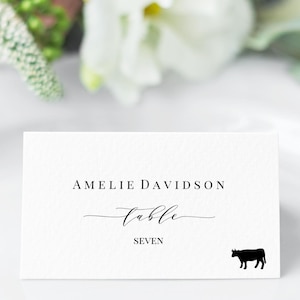 Könnte beinhalten: Eine weiße rechteckige Tischkarte mit dem schwarzen Aufdruck AMELIE DAVIDSON, dem Wort table in Schreibschrift, dem Wort SEVEN und einer kleinen schwarzen Kuh-Silhouette in der unteren rechten Ecke auf einer strukturierten Papieroberfläche.