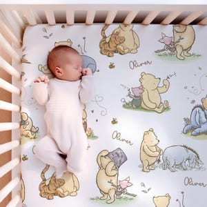 Puede incluir: Un bebé con un mono rosa claro descansa sobre una sábana de cuna con temática de Winnie the Pooh. La sábana presenta ilustraciones de Pooh, Piglet, Tigger y Eeyore, junto con el nombre "Oliver" en cursiva. Abejas y detalles florales completan el diseño.