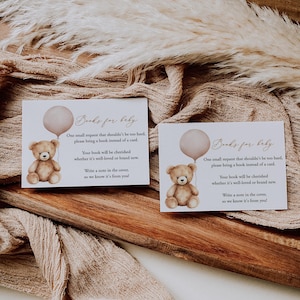 May include: Two baby shower invitation cards with the text "Books for baby". Each card features a watercolor illustration of a teddy bear holding a balloon. The cards are on a wooden surface with beige fabric and dried plants.