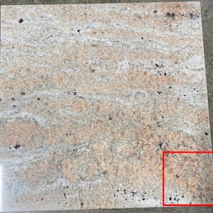 May include: A close-up of a beige and brown granite tile with a swirling pattern. The tile has a speckled appearance with small dark brown flecks throughout.