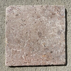 May include: A square tile made of natural stone with a light brown and beige color. The tile has a rough, textured surface with visible grains and small holes.