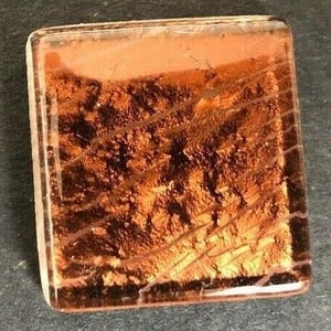 May include: A square glass tile with a copper-colored metallic finish. The tile has a textured surface with a pattern of lines and swirls.