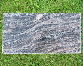 May include: A close-up of a rectangular slab of black, gray, and pink granite with a wavy pattern. The slab is sitting on a bed of green grass.