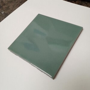 May include: A square ceramic tile with a smooth, glossy finish in a muted sage green color. The tile features straight, clean edges and a reflective surface that catches light, showcasing a uniform, solid color across its entire flat top face.