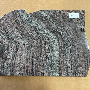 May include: A close-up of a gray, black, and pink granite slab with a wavy pattern. The slab has a rounded edge and a label that reads "M2-1".
