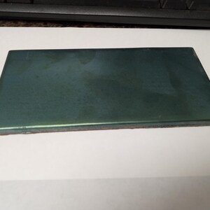 May include: A rectangular ceramic tile with a smooth, glossy finish in a deep, dark forest green color. The tile features a flat surface with slightly beveled edges and a visible reddish-brown clay base along the side profile of the object.