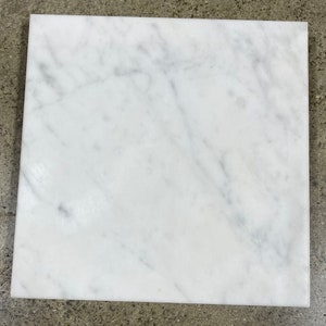 May include: A square, white marble tile with subtle gray veining. The polished surface reflects light, showcasing the natural patterns of the stone. The tile is a solid, smooth square.