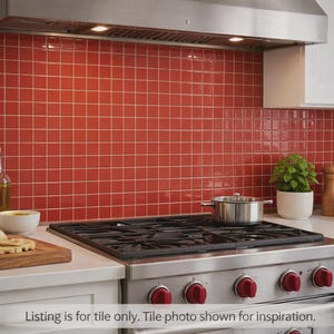 May include: A kitchen scene featuring a red tile backsplash with white grout. A stainless steel range with red knobs is in the foreground. Other elements include white cabinets, a range hood, and various kitchen items like a pot, bread, and a plant.
