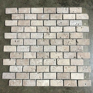 May include: A rectangular mosaic tile sheet composed of small, light beige brick-shaped tiles. The tiles are arranged in a staggered pattern, with a mesh backing visible in the upper right corner. The tiles have a textured surface and varying shades of beige and tan.