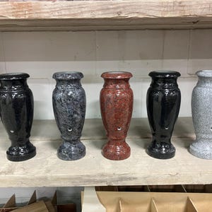 May include: Five stone vases in a row, each with a unique color and pattern. The vases are black, gray, and red, with a glossy finish. They have a classic urn shape and are displayed on a wooden shelf.