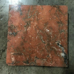 May include: A square, reddish-brown marble tile with a complex veining pattern of black, white, and gray. The polished surface reflects light, highlighting the natural variations in the stone. This tile is suitable for flooring or wall applications.