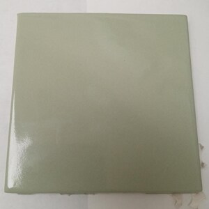 May include: A square ceramic tile with a smooth, glossy finish in a muted sage green color. The tile features straight, clean edges and a flat surface that reflects light evenly across its solid, monochromatic, and light green glazed face.