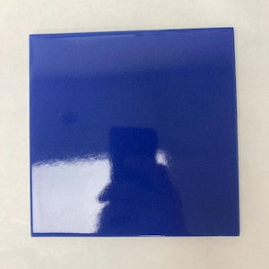 May include: A square ceramic tile with a smooth, glossy finish in a vibrant, solid royal blue color. The surface is reflective, showing a clear light reflection and a faint, distorted silhouette of the object capturing the photograph.