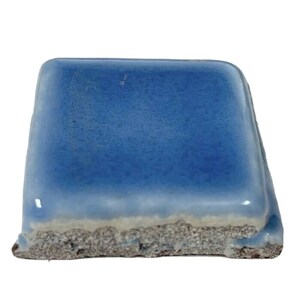 May include: A square ceramic tile with a light blue glaze. The tile has a rough, uneven edge.