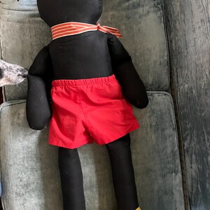 Puede incluir: Mu&ntilde;eco de peluche negro hecho a mano con pantalones cortos rojos, zapatos amarillos y una corbata a rayas. El mu&ntilde;eco tiene orejas negras grandes y est&aacute; sobre una superficie azul-verdosa. El dise&ntilde;o del mu&ntilde;eco recuerda a un personaje de dibujos animados.