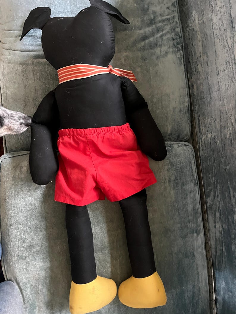 Puede incluir: Mu&ntilde;eco de peluche negro hecho a mano con pantalones cortos rojos, zapatos amarillos y una corbata a rayas. El mu&ntilde;eco tiene orejas negras grandes y est&aacute; sobre una superficie azul-verdosa. El dise&ntilde;o del mu&ntilde;eco recuerda a un personaje de dibujos animados.